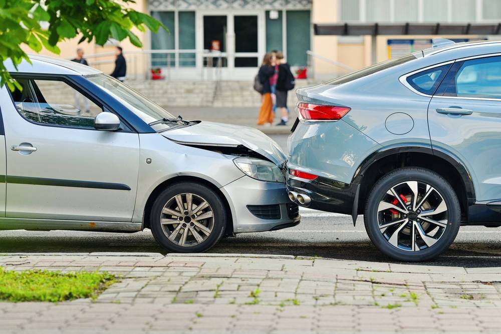 Silver SUV rear-ends a smaller silver car on a city street with pedestrians nearby