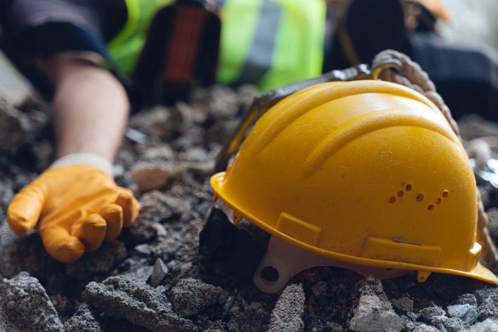 Yellow hard hat with a safety harness lying on concrete rubble beside the gloved hand of an injured construction worker